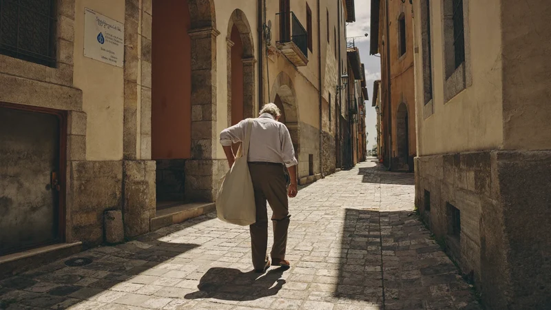 Anziano di spalle in un vicolo di pietra con borsa di stoffa